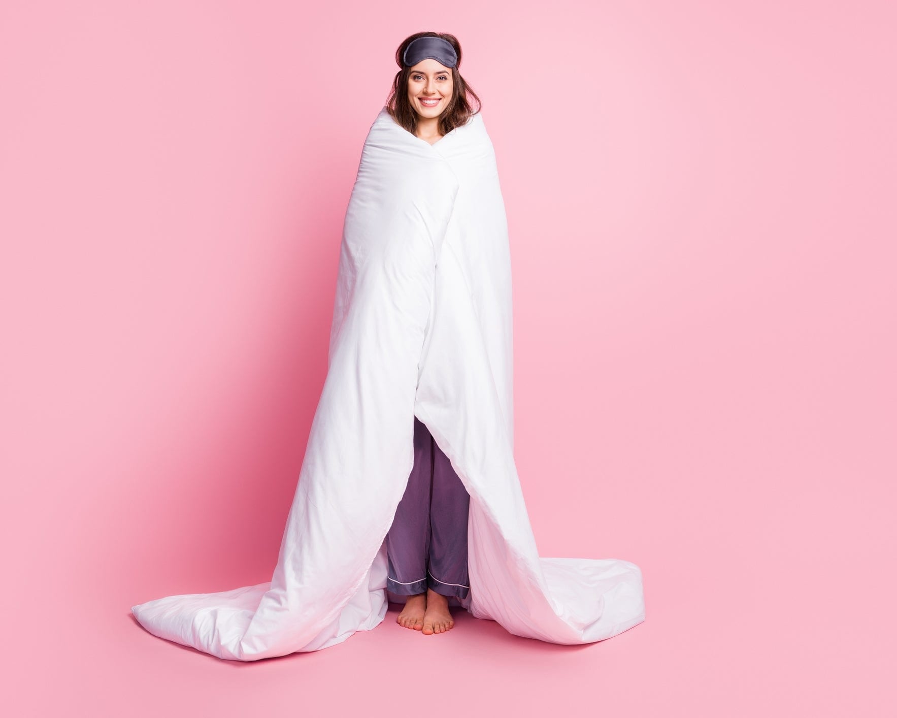 Smiling woman wrapped in a white blanket, wearing pajamas and a sleep mask against a pink background.