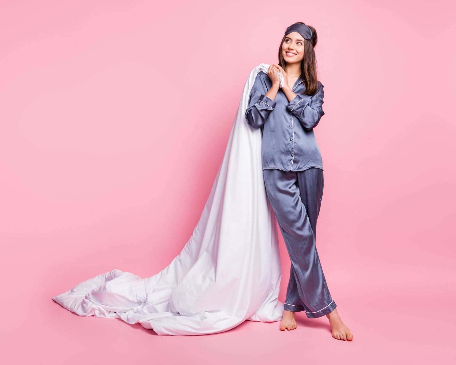 Smiling woman in gray pajamas holding a white blanket against a pink background.