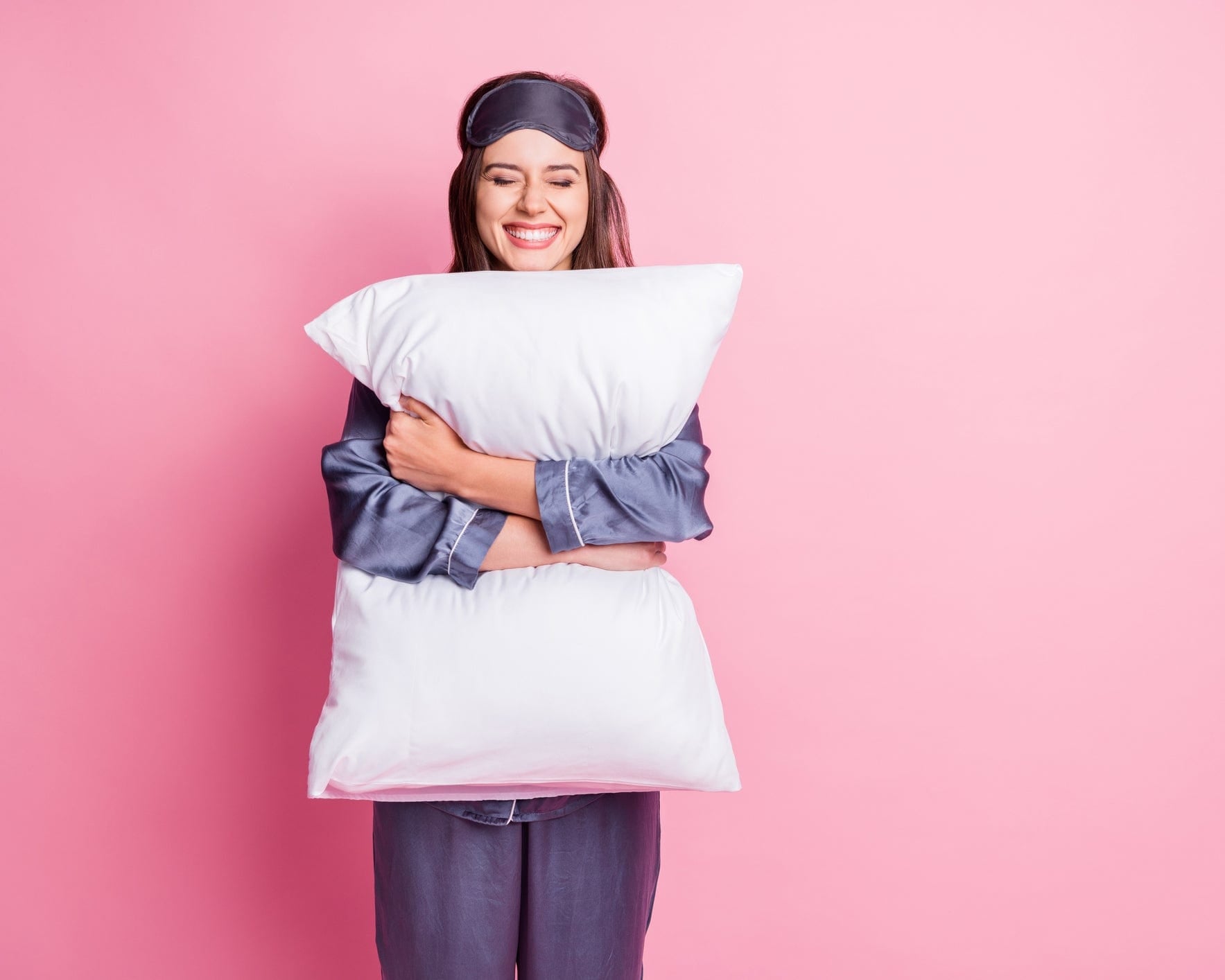 Smiling woman in sleepwear hugs a large white pillow against a pink background.