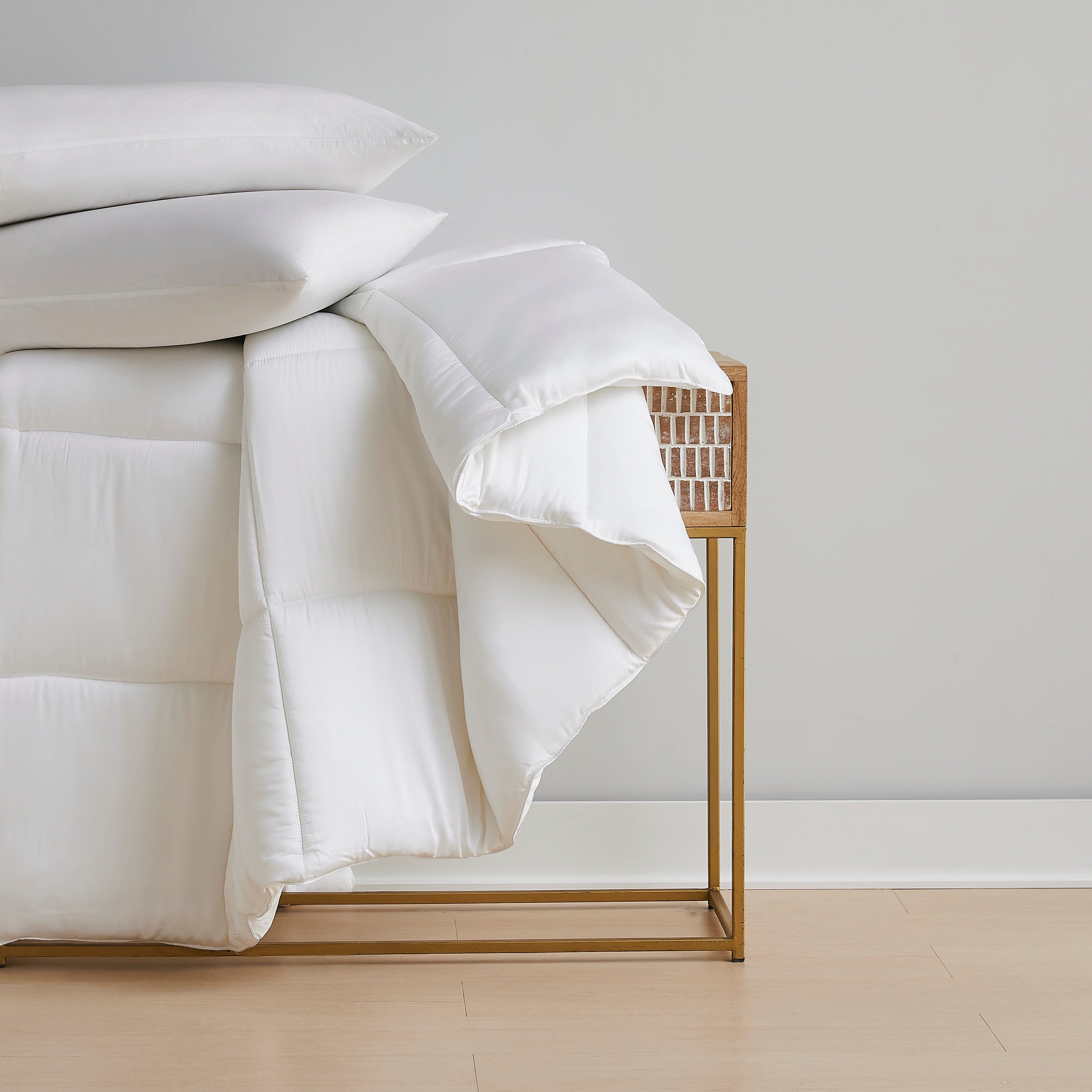 White bedding and pillows draped over a gold metal and wicker accent table.
