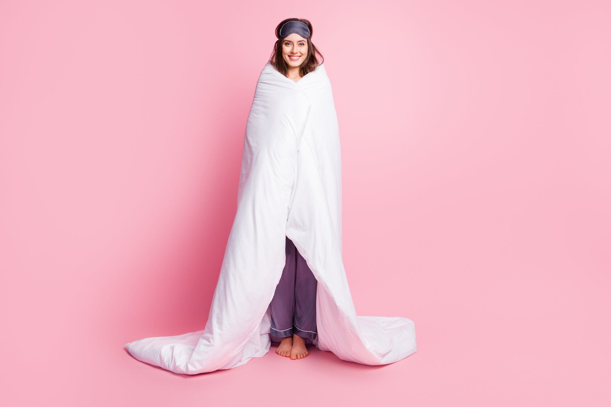 Smiling woman wrapped in a white blanket, wearing pajamas and a sleep mask against a pink background.