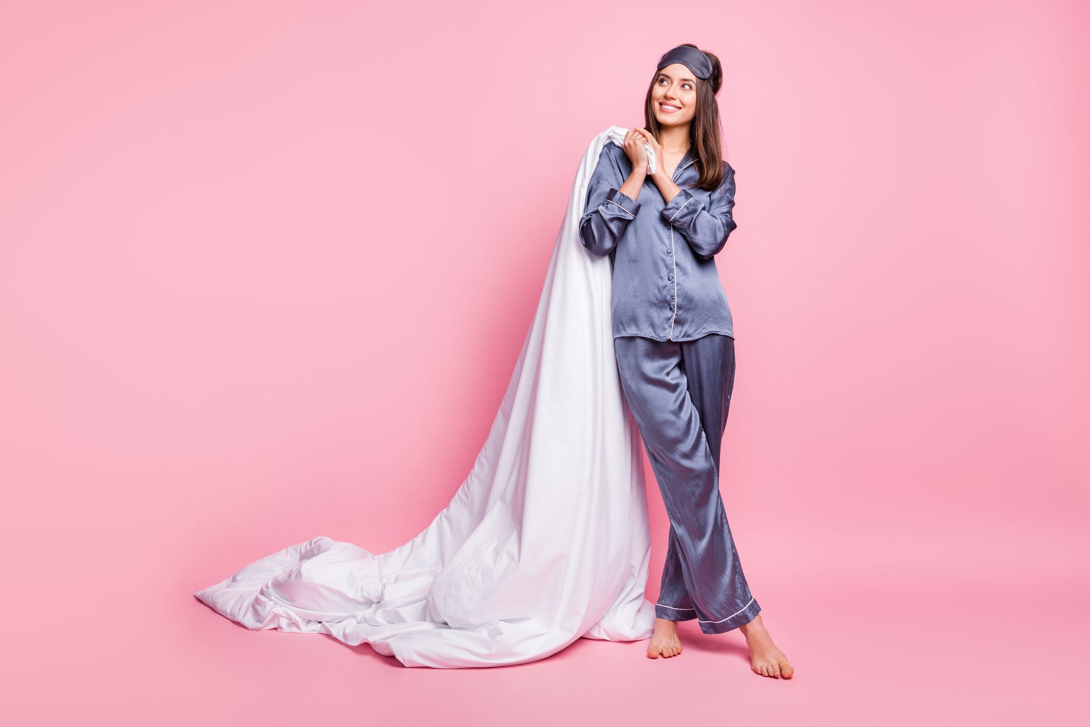 Smiling woman in gray pajamas holding a white blanket against a pink background.
