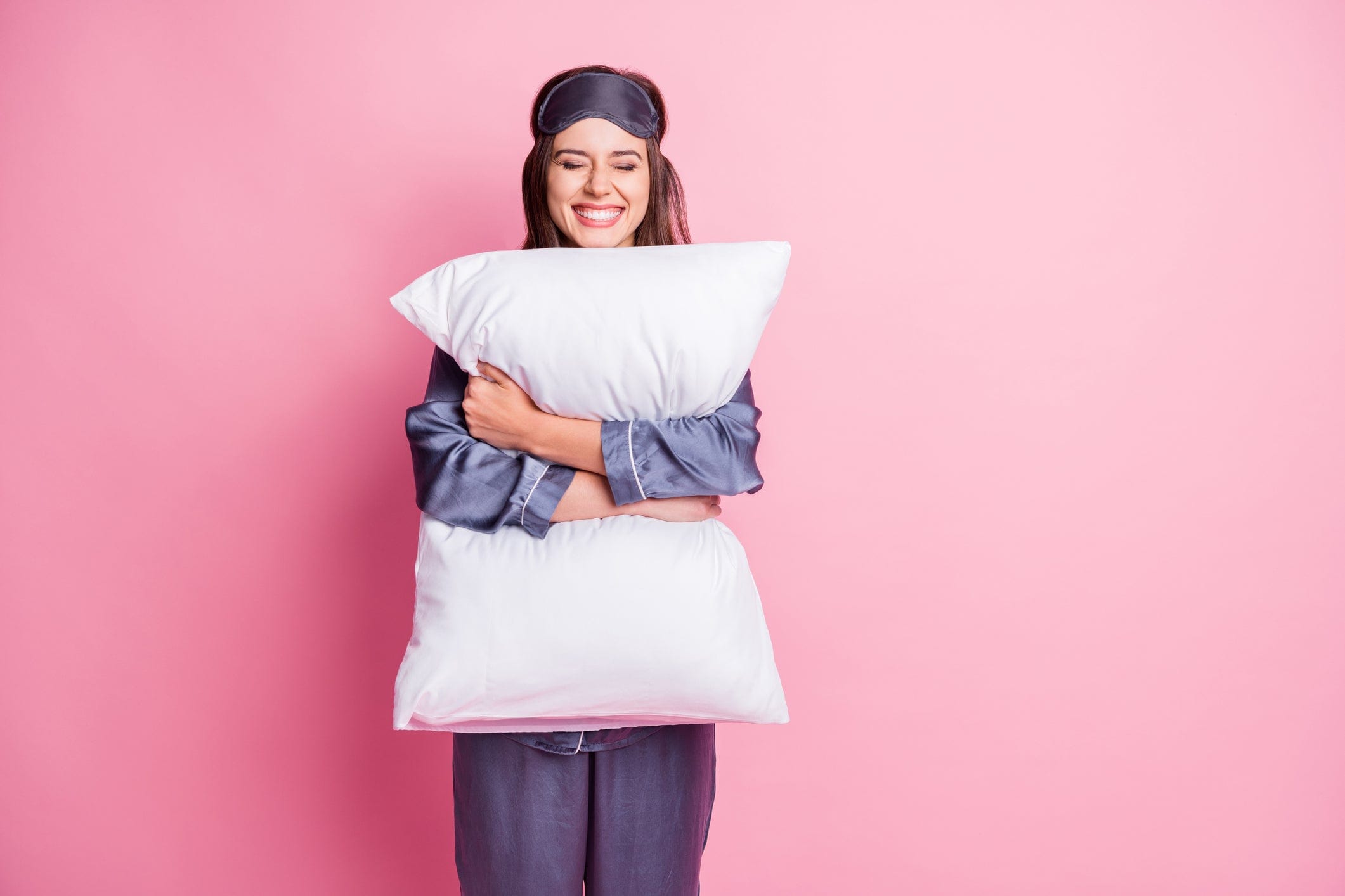 Smiling woman in sleepwear hugs a large white pillow against a pink background.